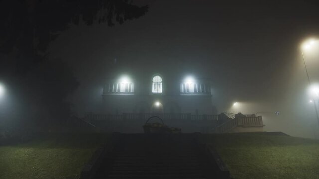 Mysterious night view of beautiful abandoned haunted house with lights inside. Cinematic horror scene. Foggy evening. Rainy weather.