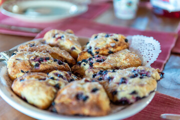 A white porcelain plate with a white paper doily and blueberry scones covered in white sweet icing. The homemade sweets are placed in the middle of brown wooden dining with burgundy placemats.  