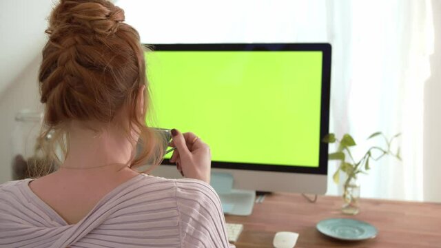 Concept for drink, tea or coffee. Young women sitting in front of computer with green screen mock-up. Girl drinking Coffee or tea. Over shoulder camera shot 4k video footage.