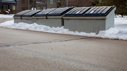 Three large metal garbage bin sits on the side of a street in a condo corporation for residents to dispose of their garbage. The bin is old and rusty, and in need of a better waste management system