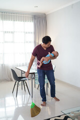 portrait of asian father cleaning the floor using broom while carrying his infant baby