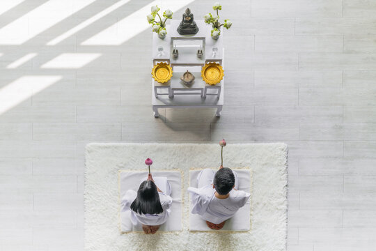 asian couple wearing white clothing worship and praying to buddha statue together with holding lotus flowers in hands