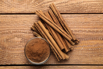 Aromatic cinnamon powder in bowl near sticks on wooden table, flat lay