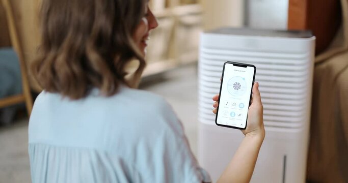 Woman controlling with a smartphone portable air conditioner, holding phone with launched program for smart home appliances