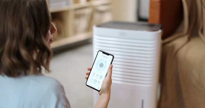 Woman controlling with a smartphone portable air conditioner, holding phone with launched program for smart home appliances