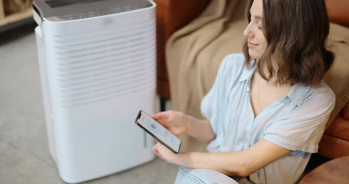 Woman controlling with a smartphone portable air conditioner, holding phone with launched program for smart home appliances