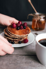 Hands hold white plate with pancakes, berries, honey, coffee cup on wooden table