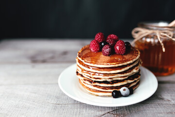 Juicy pancakes with berries and honey on a white plate, spoon, jar, wooden table