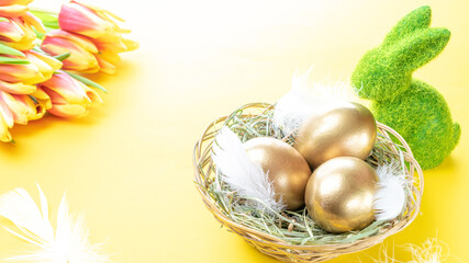 Golden eggs in basket with spring tulips, white feathers on pastel yellow background in Happy Easter decoration. Foil minimalist egg design, modern design.