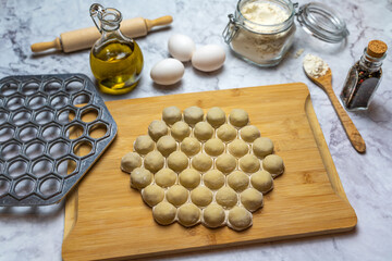 Raw meat dumplings on a cutting board. There are flour, eggs, rolling pin, dumpling maker, olive oil and pepper. 