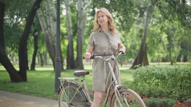 Wide shot of cheerful blond middle aged woman riding bicycle in sunny summer park and stopping. Portrait of excited happy Caucasian lady enjoying bike riding outdoors. Hobby and healthy lifestyle.