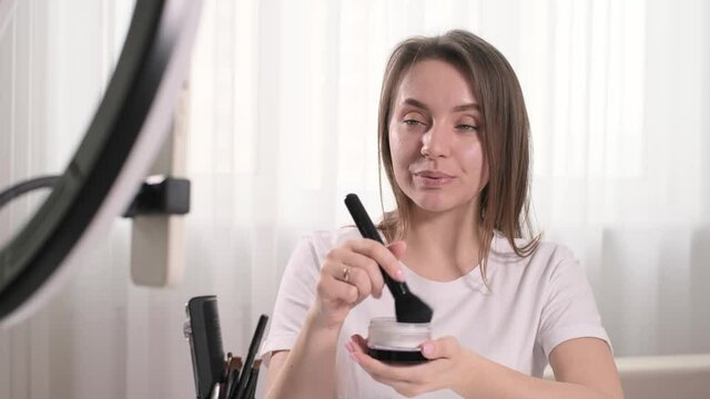 Woman blogger doing make-up at home, apply decorative cosmetics, powder. Beautiful young female using brush near the mirror in the living room, self care and product presentation for users online 