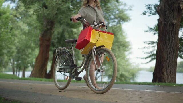 Wide shot portrait of confident positive middle aged woman walking with shopping bags hanging on bicycle steering wheel. Happy smiling shopper strolling with bike outdoors on summer day.