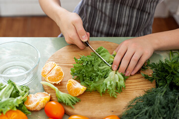 A teenage boy prepares a fruit salad on a recipe from the Internet. The boy cuts lettuce on the kitchen table, in front of him is a laptop. The child is engaged in household chores.