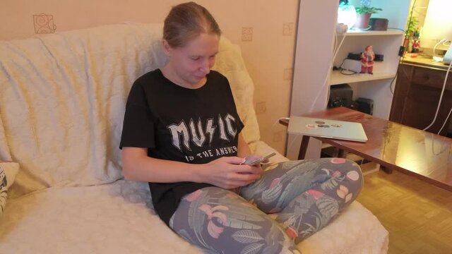 A young girl sits on the couch in a room of her house and reads mail on the phone, then delightedly rejoices and laughs. Woman Swipes the chat and receives a funny sms. 