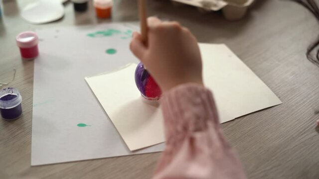 A child's hand with a paintbrush paints an Easter egg on white paper on a wooden table. Easter holiday for children and a painted egg.
