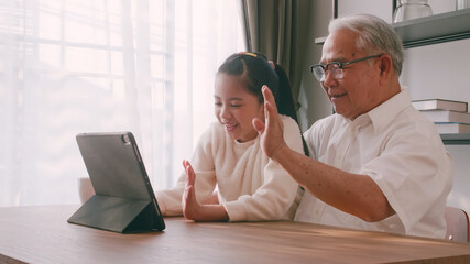 An Asian grandfather and granddaughter spending time together in the living room. Happy senior man with little girl using a tablet on the table at home.