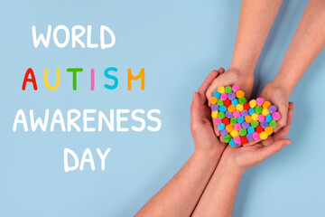 World Autism Awareness Day concept - autistic child's hands supported by mother holding multicolored heart on blue background. Autism spectrum disorder and child mental health concept, selective focus