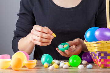 Fototapeta na wymiar A caucasian woman is filling plastic easter egg shells with crisp sugar coated chocolate eggs in preparation for the easter celebration. She puts the completed ones in basket ready for egg hunt.