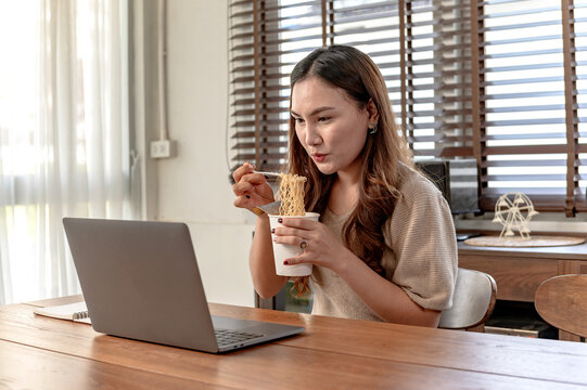 Asian Businesswoman Working Alone Online At Home. Woman Eating Cup Noodle And Busy.