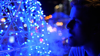 Bokeh view of a stylish young handsome man smoking electronic cigarette and breathing out smoke thru nose in city at night in summer