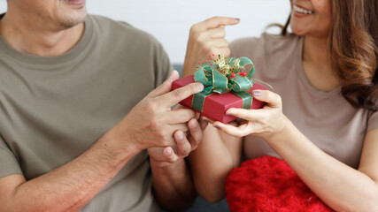 Portrait of cute smiling middle-aged Asian lover couple wearing eyeglasses in casual clothes sitting on sofa indoor. Husband surprising his wife with a red and green gift box on Christmas eve holiday