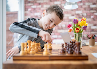 Cute young happy boy chess genius concentrating playing game of strategy moving piece sitting at wooden table with chessboard on bright sunny summer day