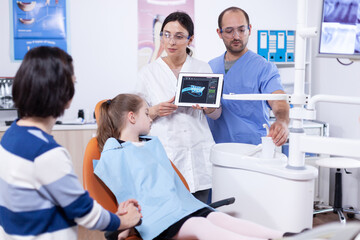 Obraz premium Dentist in dentistiry office showing tablet pc with jaw radiography to parent and little girl explaining treatment. Stomatologist explaining teeth diagnosis to mother of child in health clinic holding