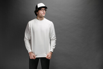Man wearing blank white sweatshirt and empty baseball cap standing over gray background. Sweatshirt...