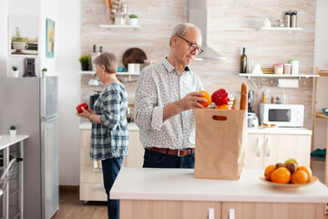 Elderly husband and wife taking out vegetables from grocery paper bag after arriving from supermarket. Cheerful happy family healthy lifestyle putting fresh fruits and groceries in refrigerator