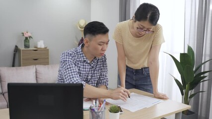 asian wife walking up to her husband to discuss about bank loans with their finger pointing at the data on the document at home. - Powered by Adobe