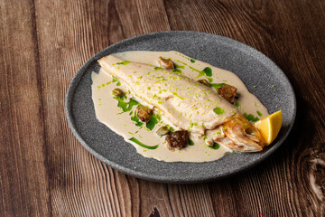 Baked fish with cream sauce and a slice of lemon isolated on a grey round plate. Close-up of a dish on wooden background. Horizontal orientation, copy space. Restaurant menu concept.