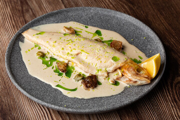 Baked fish with cream sauce and a slice of lemon isolated on a grey round plate. Close-up of a dish on wooden background. Horizontal orientation, copy space. Restaurant menu concept.