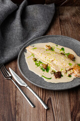 Baked fish with cream sauce and a slice of lemon isolated on a grey round plate. Close-up of a dish on wooden background with grey linen napkin and silver cutlery by side. Restaurant menu concept.