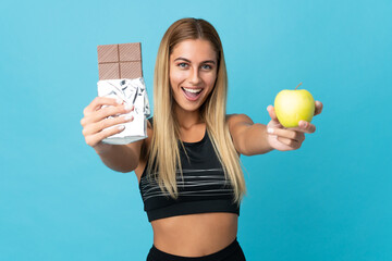 Young blonde woman isolated on blue background taking a chocolate tablet in one hand and an apple in the other