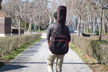 Latin young man backwards carrying a guitar in a guitar case on a city street. University campus.