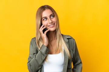 Young blonde woman isolated on yellow background keeping a conversation with the mobile phone with someone