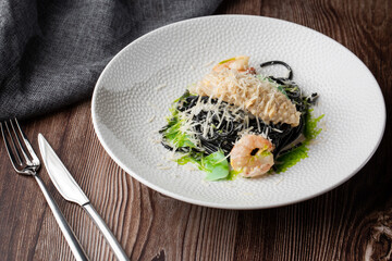 Black spaghetti. Black seafood pasta with shrimps and parmesan on white plate isolated on wooden background. Silver cutlery and grey linen napkin by side. Mediterranean gourmet food.