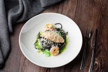 Black spaghetti. Black seafood pasta with shrimps and parmesan on white plate isolated on wooden background. Silver cutlery and grey linen napkin by side. Mediterranean gourmet food.