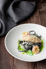 Black spaghetti. Black seafood pasta with shrimps and parmesan on white plate isolated on wooden background with grey linen napkin by side. Vertical orientation. Mediterranean gourmet food.