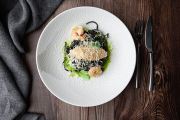 Black spaghetti. Black seafood pasta with shrimps and parmesan on white plate isolated on wooden background. Silver cutlery and grey linen napkin by side. Mediterranean gourmet food. Top view.