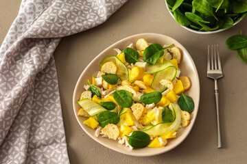 Plate with salad and fork close-up on a brown background, salad of cucumber and chicken breast, mozzarella and croutons with sweet mango pieces and spinach