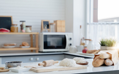Blur background of a wooden rolling pin and 2 dough on wooden board tray with three loaves of bread in a paper bag place on the table in a small modern kitchen with the window
