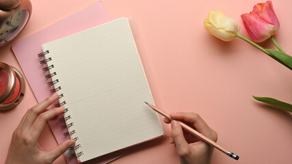 Female hand with pencil writing on blank notebook on feminine workspace with flower