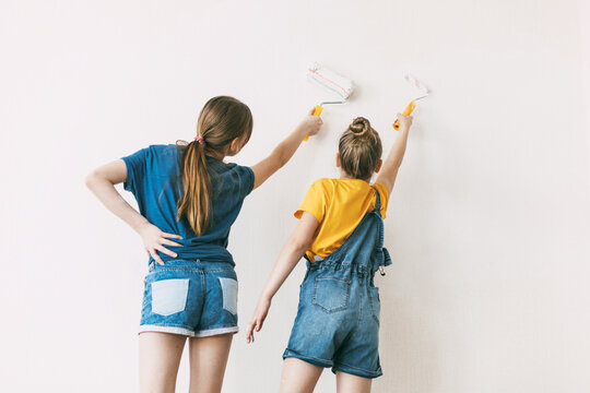 Two sisters are painting the walls of the house white, in denim overalls and a bright yellow T-shirt, home renovation