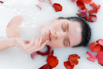 Fototapeta na wymiar Close up female face in the milk bath with soft white glowing and rose petals. Copyspace for advertising. Beauty, fashion, style, bodycare concept. Attractive caucasian model in milky colored foam.