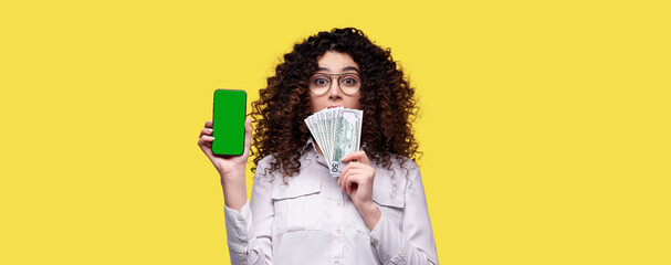 Surprised woman in glasses holds stack of dollars and smartphone with blank green screen over yellow isolated background. Concept of online casino, bet, games