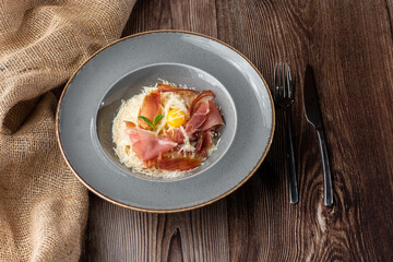 Pasta Carbonara. Spaghetti with bacon, parmesan, cream sauce and egg yolk. Isolated on a grey round plate with silver cutlery and sacking on wooden background. Mediterranean gourmet food.
