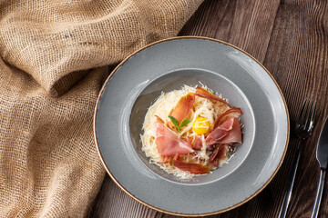 Pasta Carbonara. Spaghetti with bacon, parmesan, cream sauce and egg yolk. Isolated on a grey round plate with silver cutlery and sacking on wooden background. Mediterranean gourmet food.