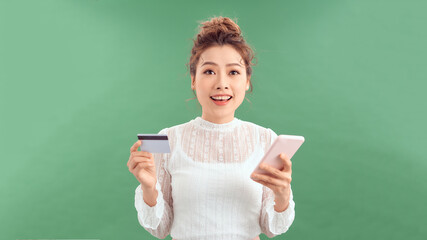 Happy young woman standing isolated over green background, holding mobile phone, showing plastic credit card